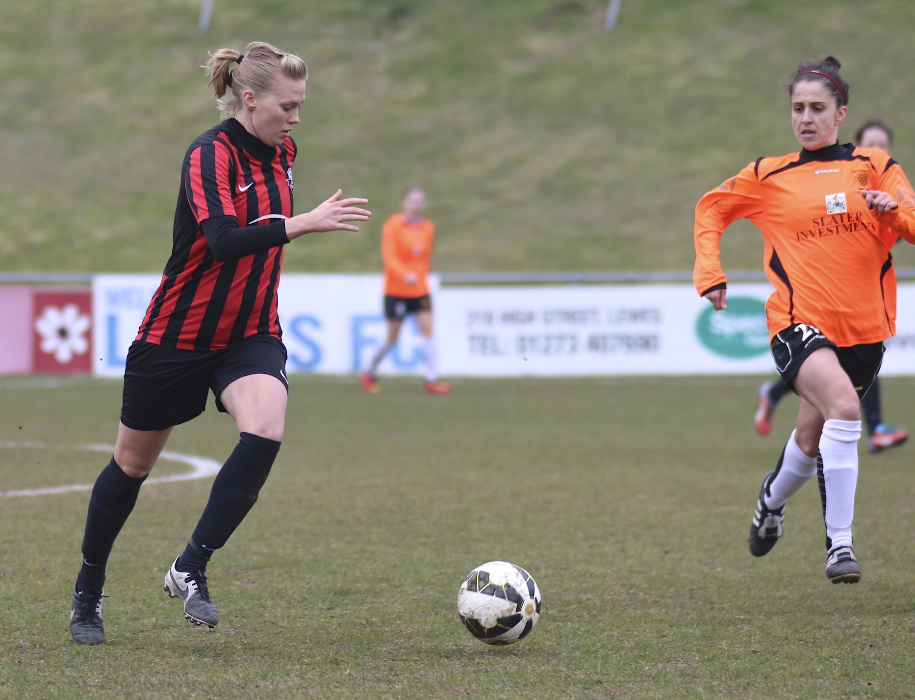 two women play soccer on the field with other people watching