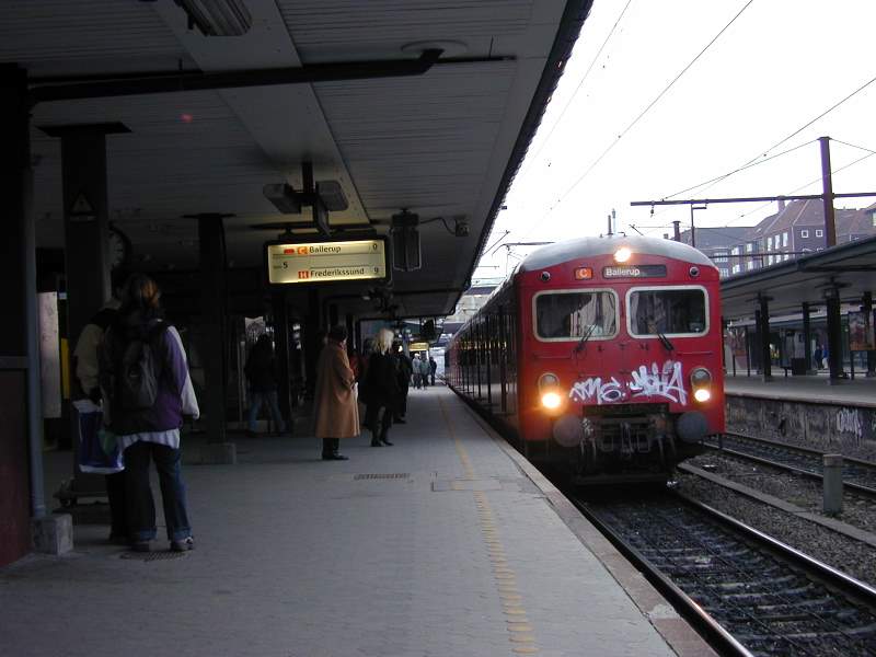 a red train pulled up to a train station with people walking near by