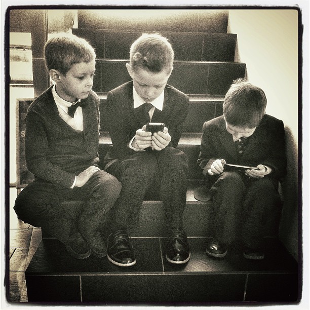 three boys dressed in suits sit on a set of stairs looking at their cellphones