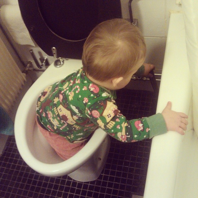 small child in green shirt standing at edge of toilet with arms spread