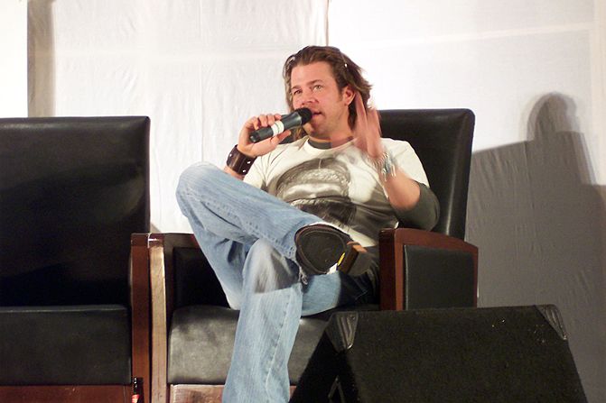 a man in jeans, a white shirt and black shoes sitting in a chair with a microphone