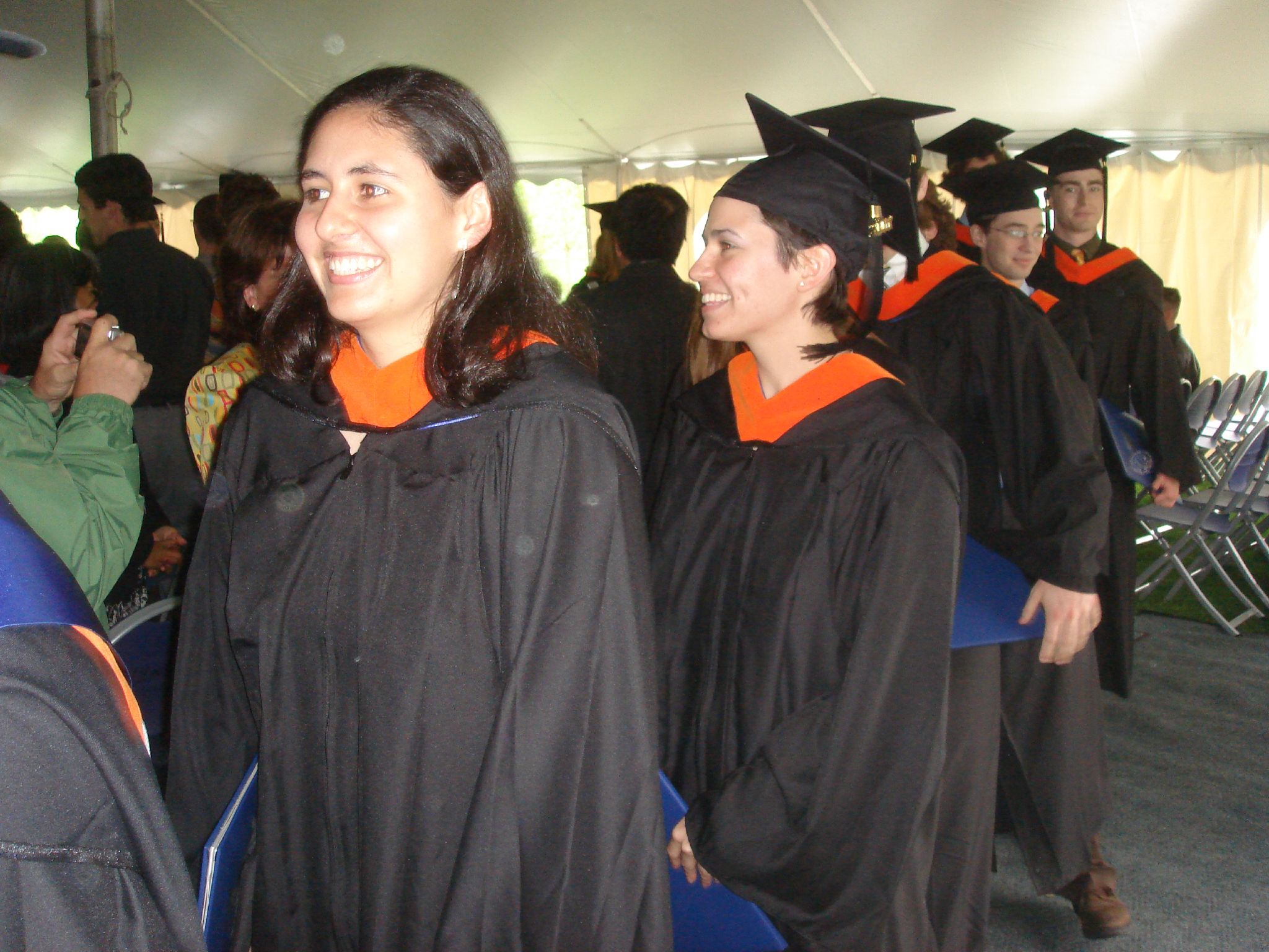 two girls in black graduation gowns and orange accents standing together with other graduates