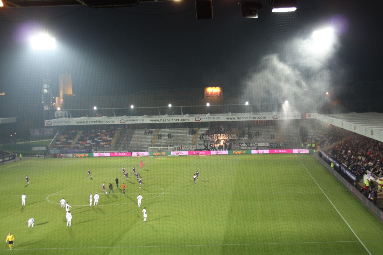 a soccer field with smoke pouring from the stands and onlookers watching