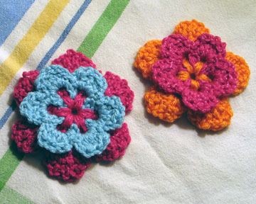 two small crocheted flowers, one in red, blue and the other in pink