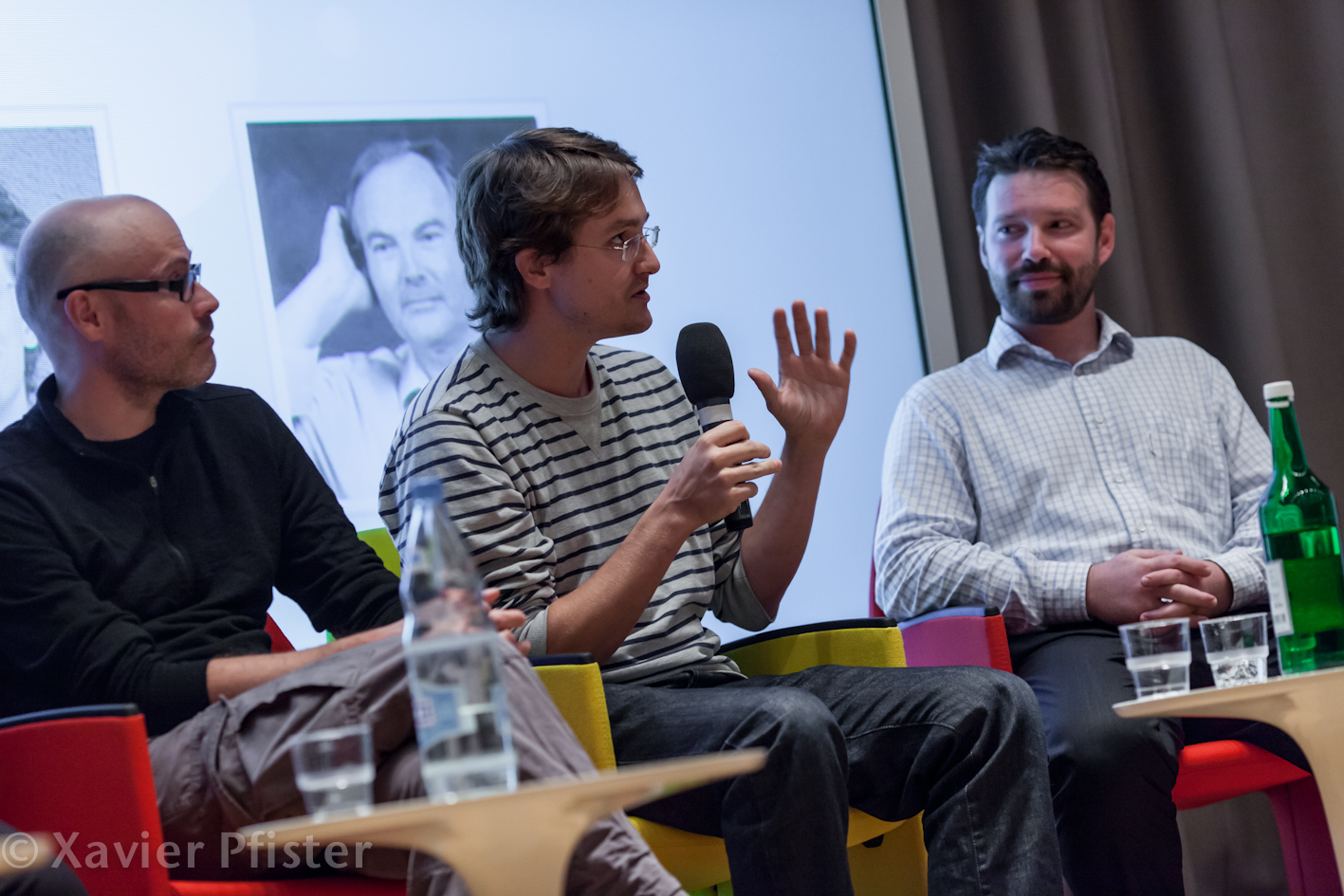 three men are sitting on chairs in front of a projector screen while talking to each other