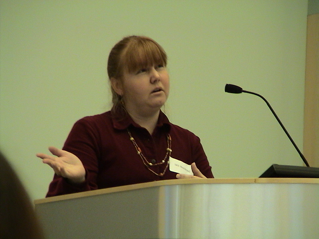 woman with red hair and gold necklace sitting in front of a podium and microphone