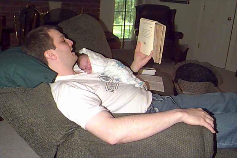 a man is sitting in his chair reading a book while holding his baby