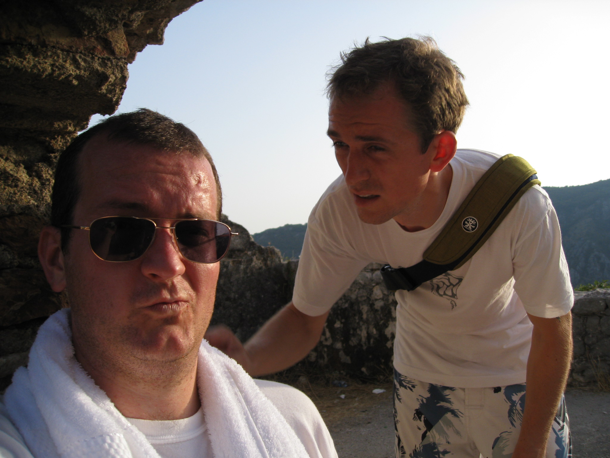 a man in a towel and sunglasses holds out his camera to an older man in a white shirt