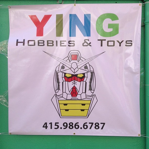 a sign for ying hobbies & toys hangs on a green wall