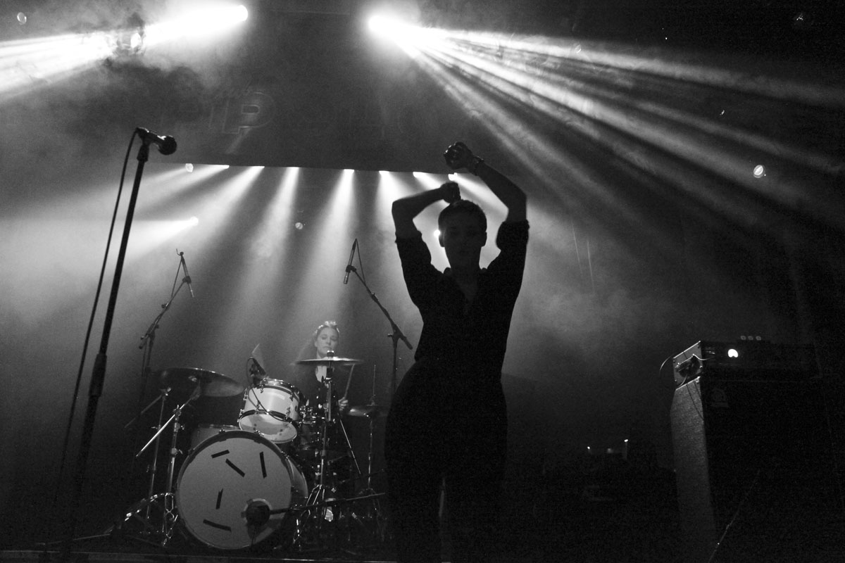 black and white pograph of person holding up his arm in the air in front of drum set with lights behind him