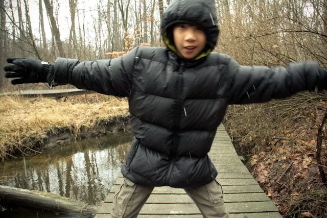 a boy in a coat and hat on a boardwalk crossing a creek