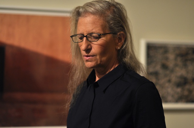 an older woman with glasses stands next to some wood paneling