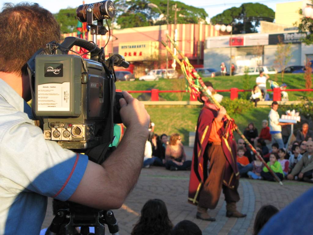 a crowd watches as a man with an elaborately adorned sash is filmed by a camera