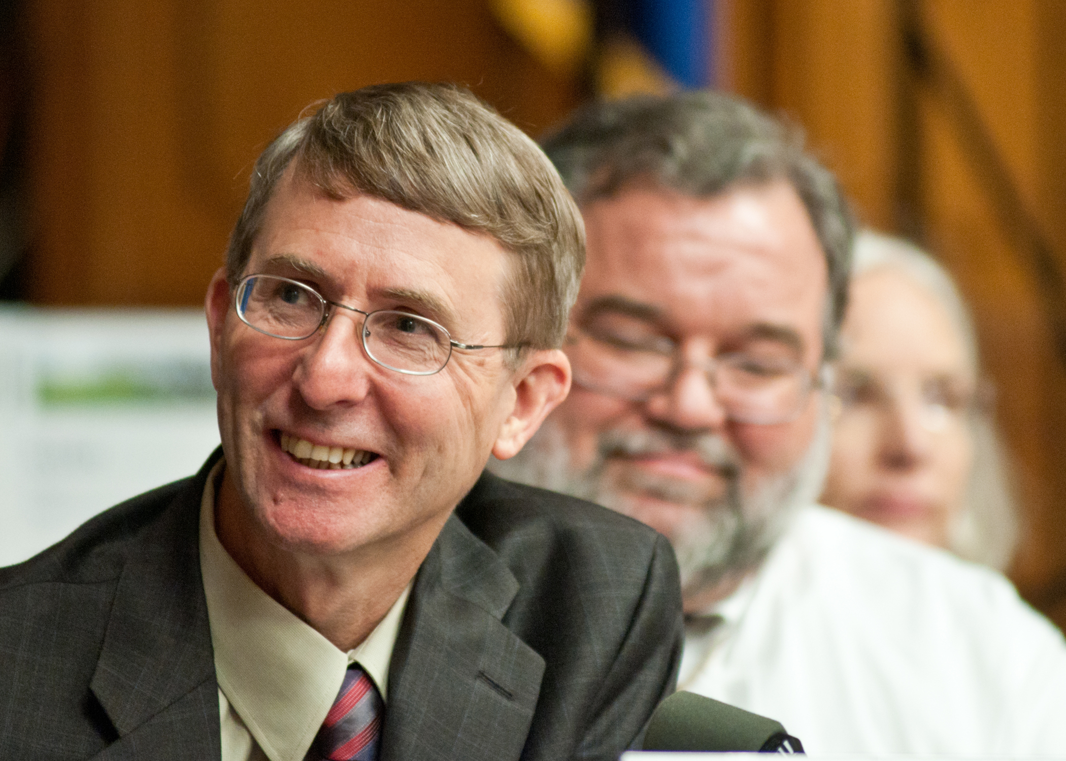 an older man in a gray suit smiling while two others watch