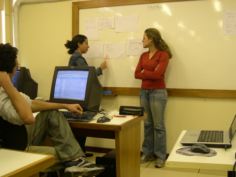 woman is talking in front of whiteboard as another person uses a laptop