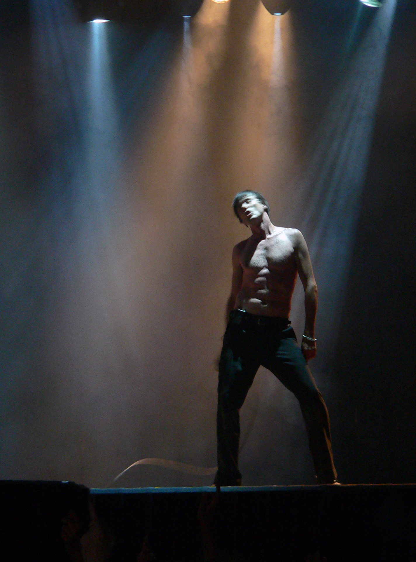 shirtless man stands on stage with light beams