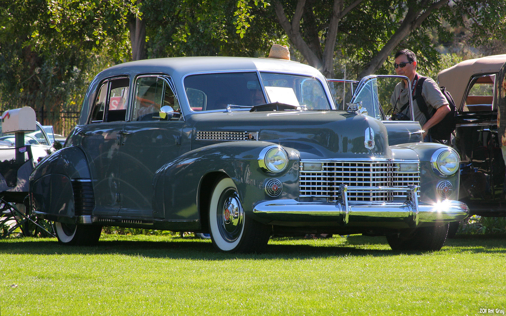 an old - fashioned car is parked in the park while a man in sunglasses stands next to it