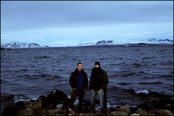 two men are standing by the water with snow covered mountains behind them