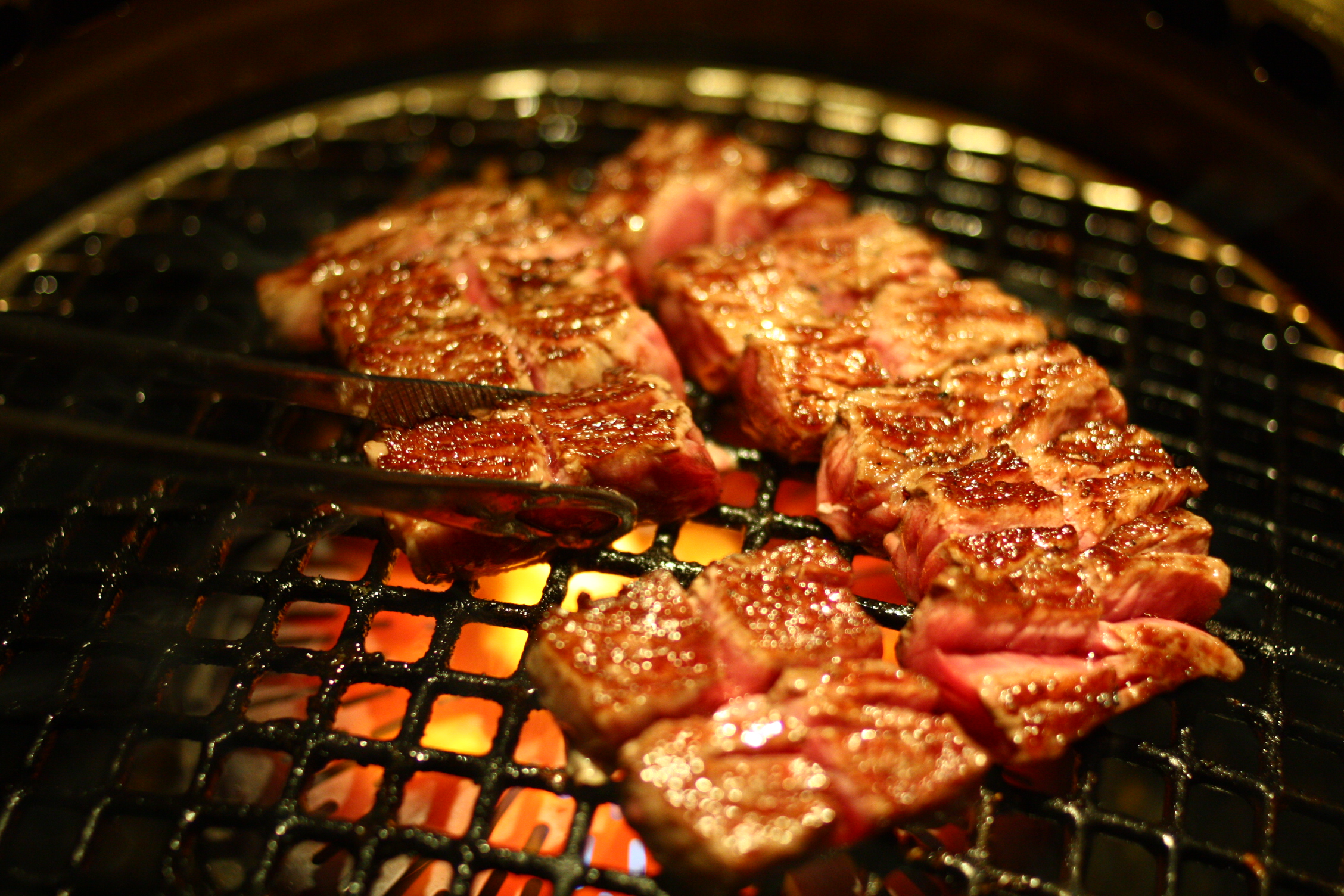 steak cooking on an open barbecue grill while someone uses tongs to cut it