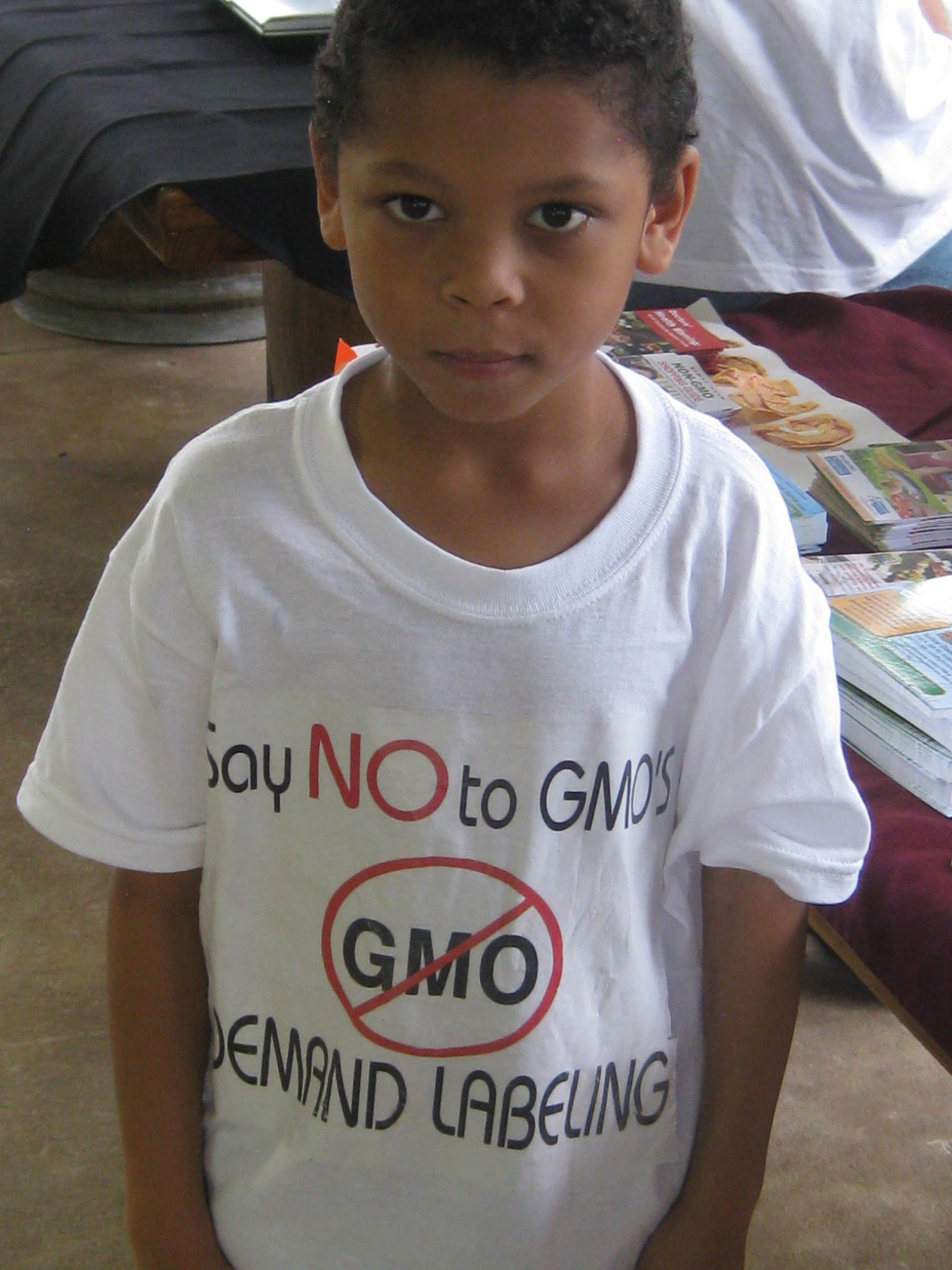 a boy is standing wearing a white shirt that reads, say no to gmo and stands in a room