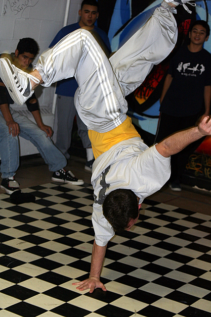 a man in grey shirt doing a flip trick on a checkered floor
