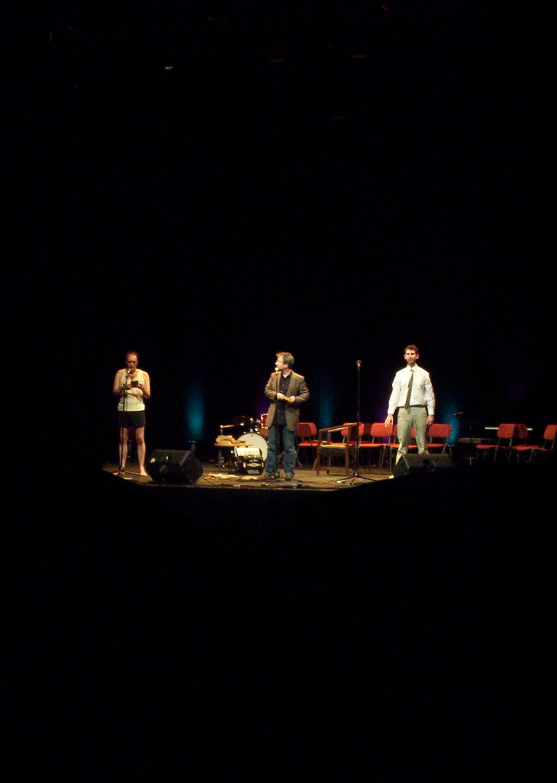 three men stand in front of a group of empty chairs on a stage