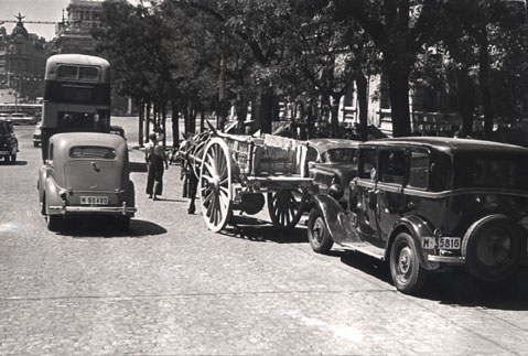 a vintage po with horses, cars and people walking on the sidewalk
