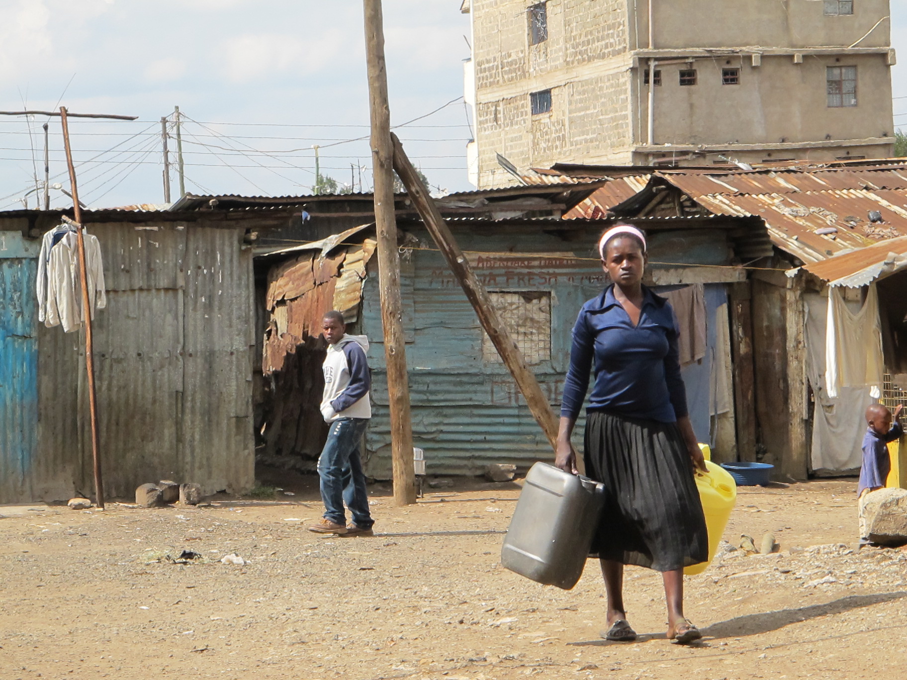 a woman carrying two containers in her hand in front of some shacks