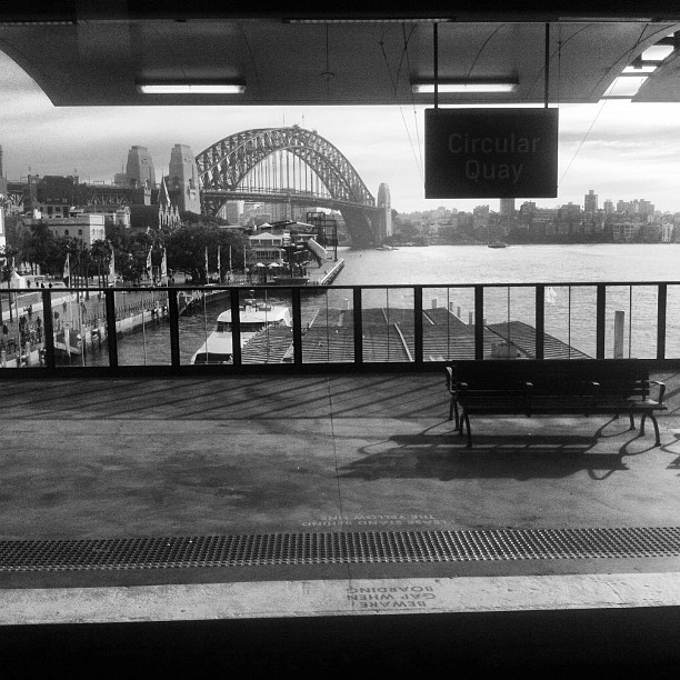 an empty bench sitting near the water with a view of a bridge in the background