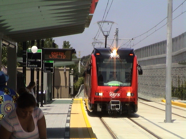 a red and black train passing by a woman walking on the sidewalk