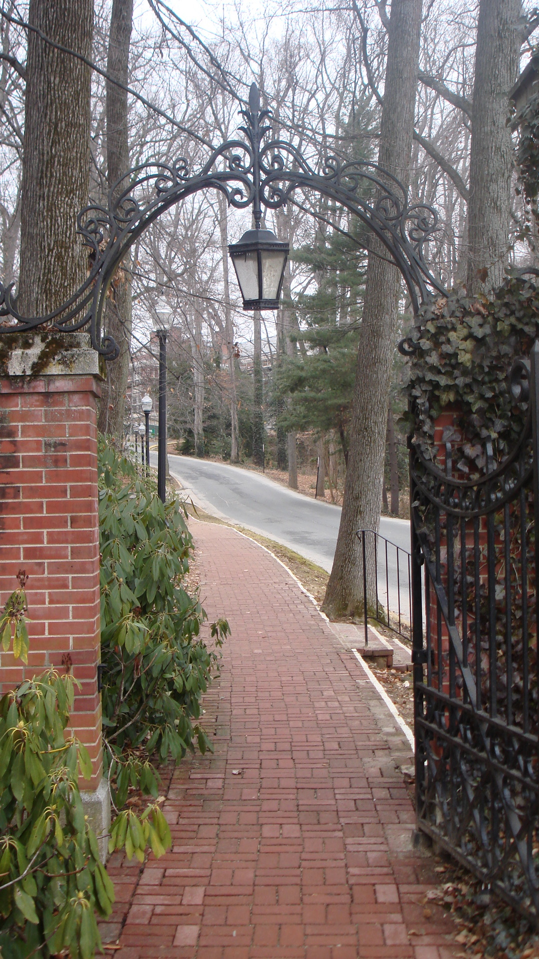 an arched iron gate leads into a path leading to a red brick path in the woods
