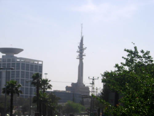 an empty street with people walking, cars driving down it and a very tall tower in the background