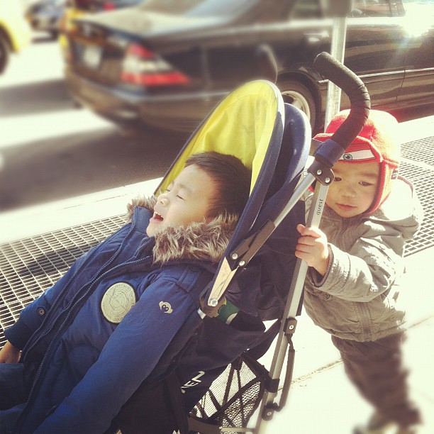 a child holding onto an attached stroller that sits on the side of a sidewalk next to another child wearing warm clothing