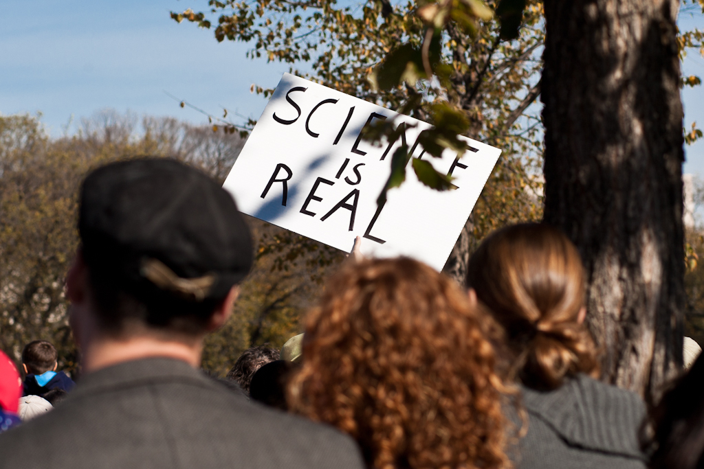 people standing in the shade outside, while a sign reads science is real