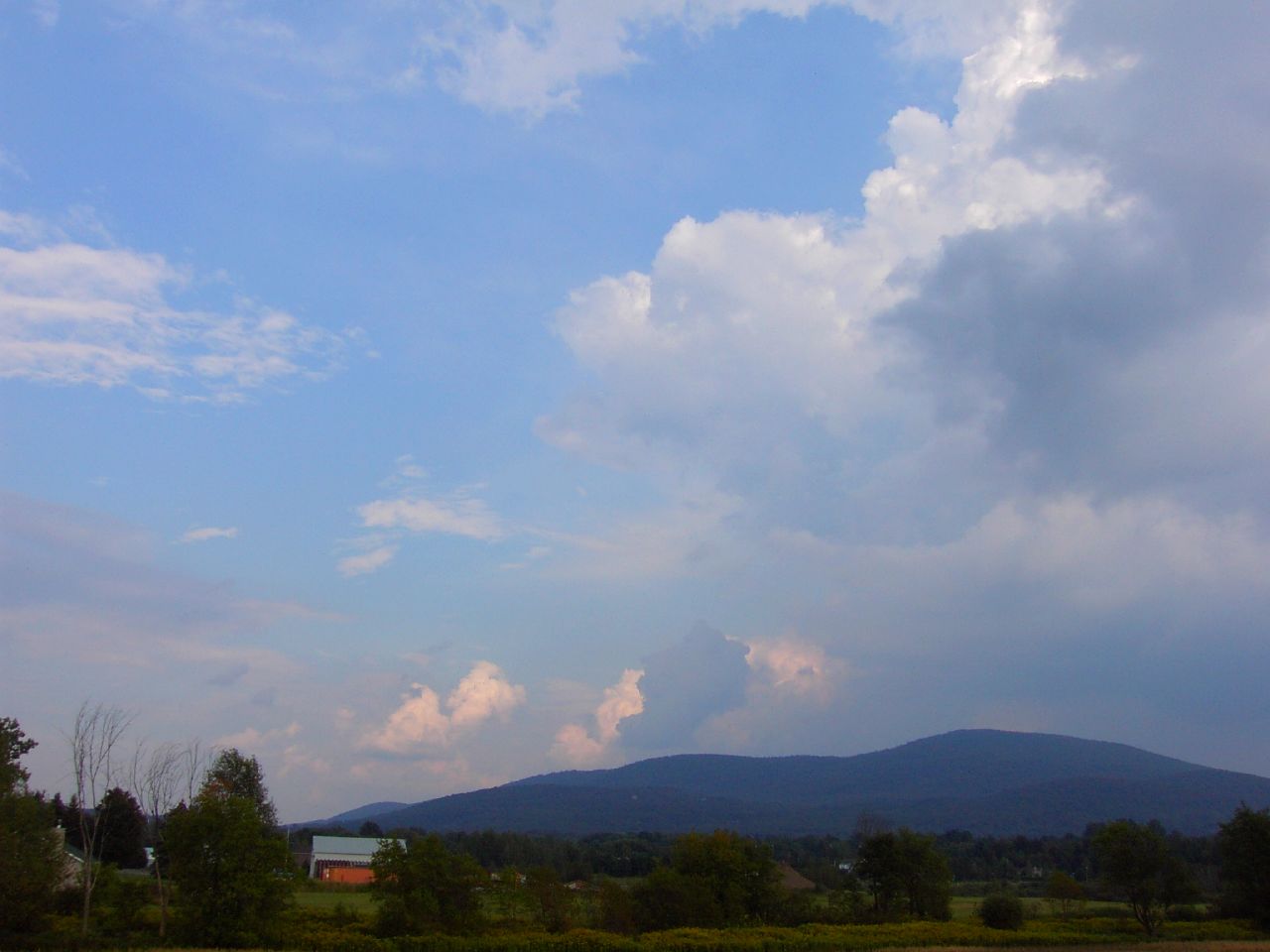 a blue sky over a green field with a farm in the foreground and some mountains in the background