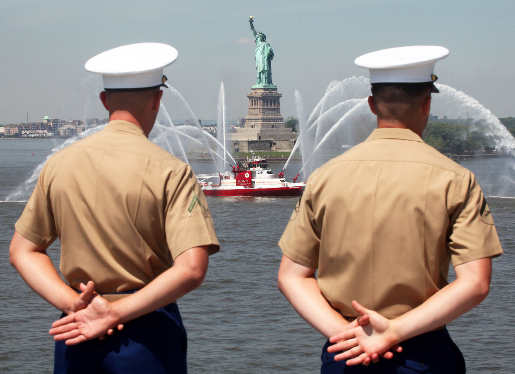 two men in tan shirts standing in front of the statue of liberty