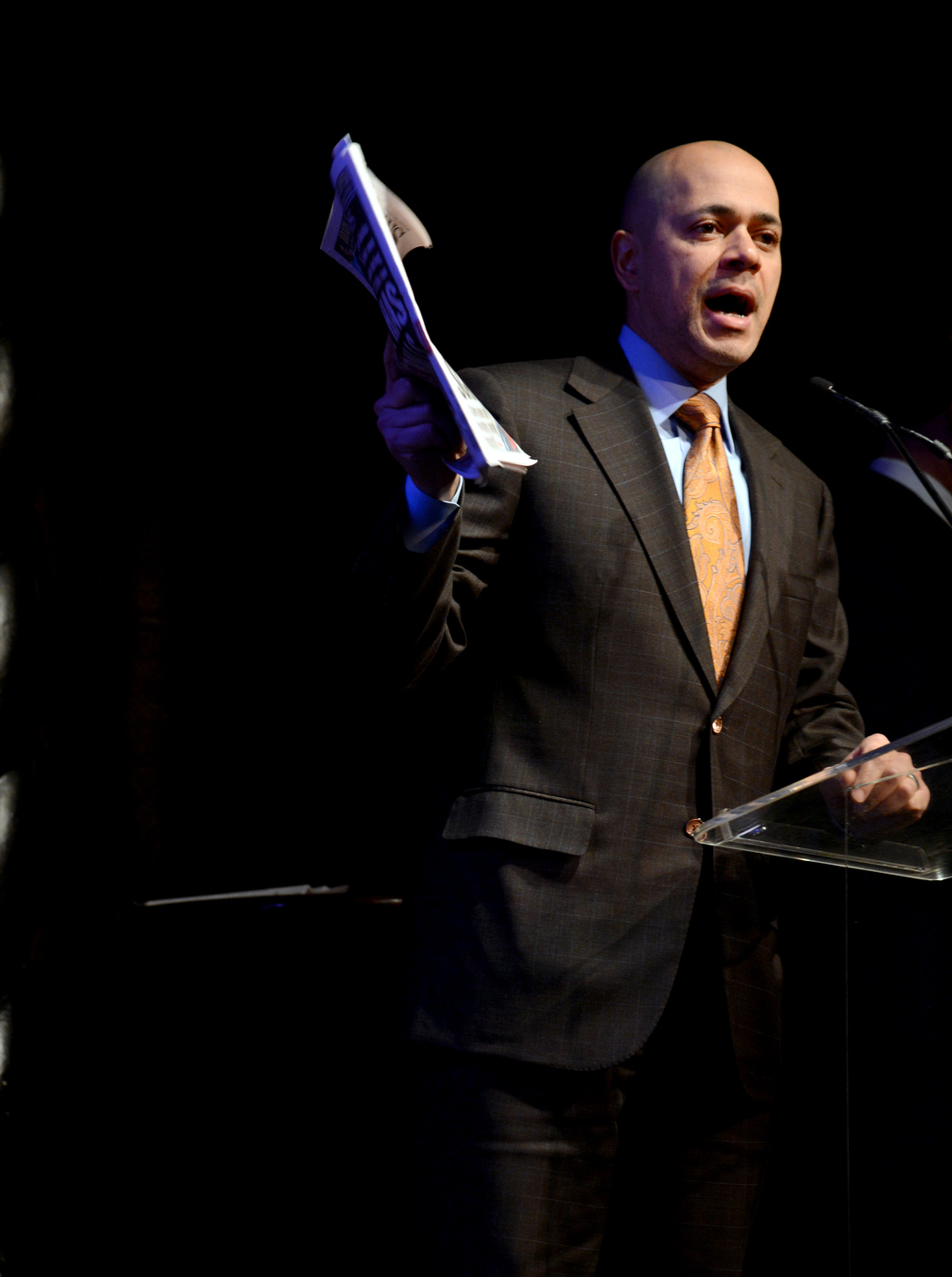 a man in suit and tie holding a newspaper on stage