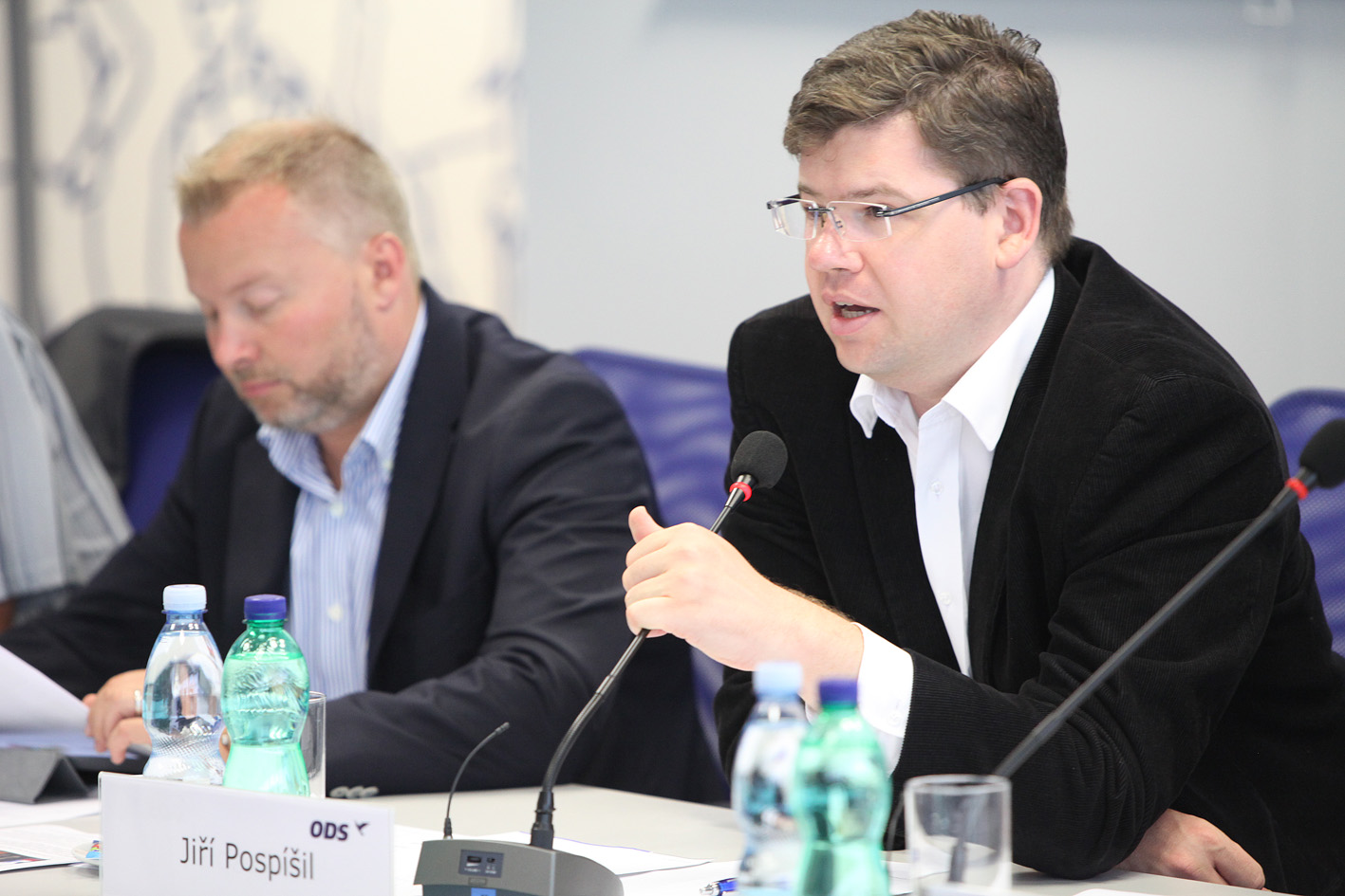 a person in a black jacket speaking at a table with bottles of water