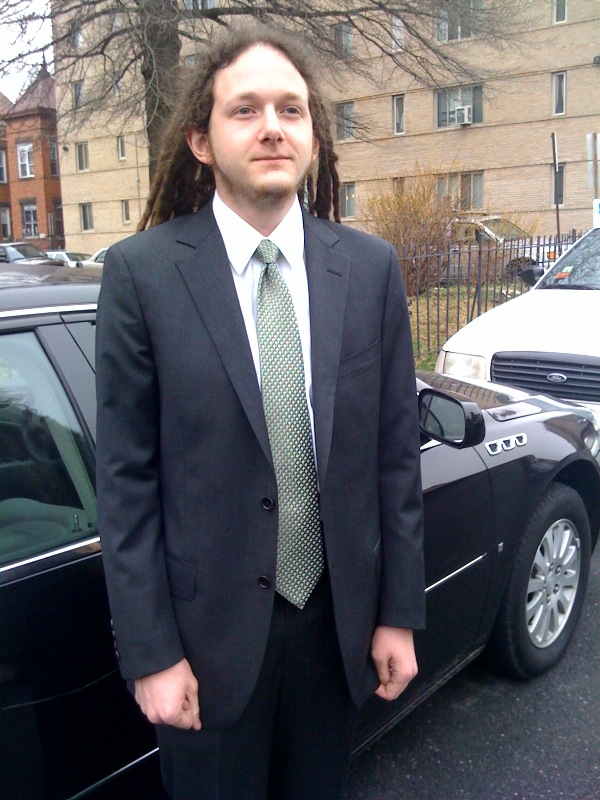 a young man dressed in business attire posing next to his car