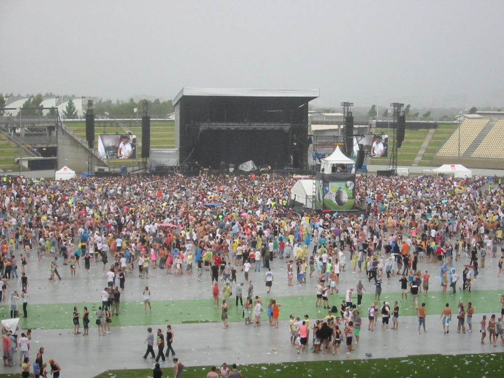 a crowd of people walking along a wet sidewalk next to a stage