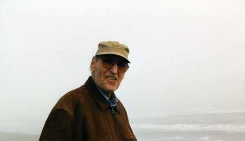 an old man wearing a hat and brown jacket with ocean waves