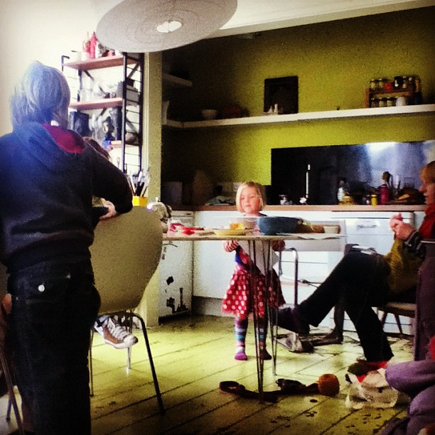 a little girl in a red dress sitting at a table with adults in a kitchen