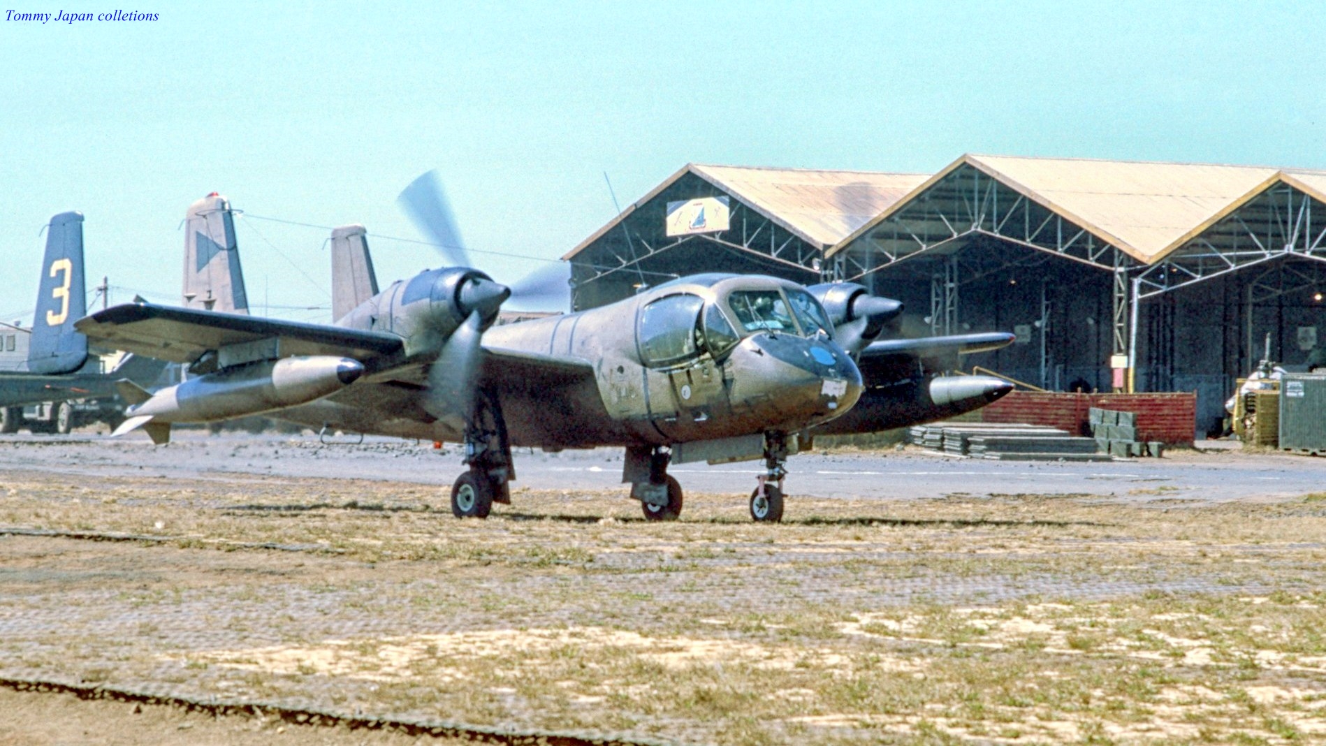 the plane sits outside in the dirt next to several hangars