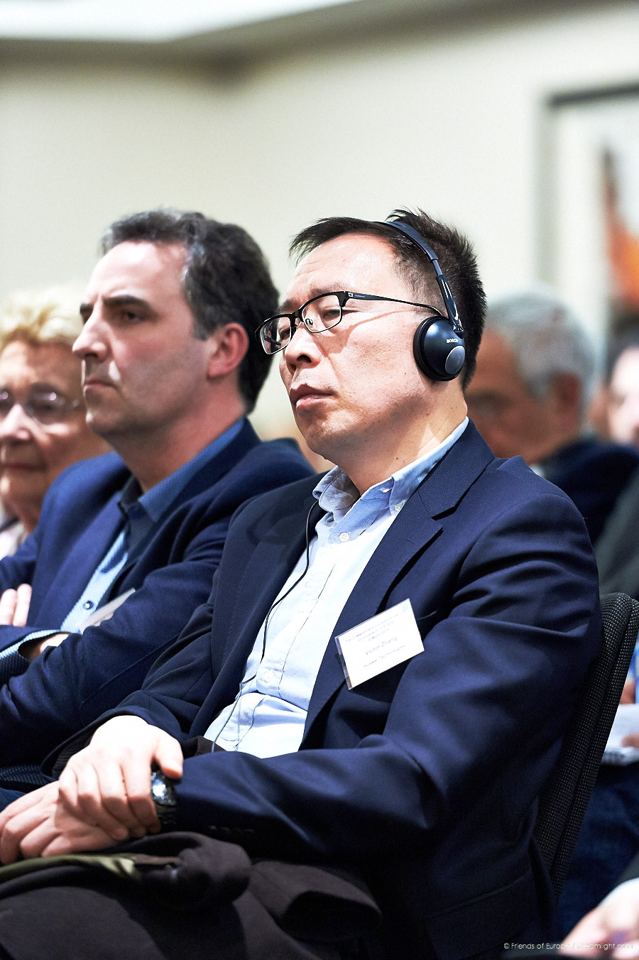 three men in business attire sit listening to someone else