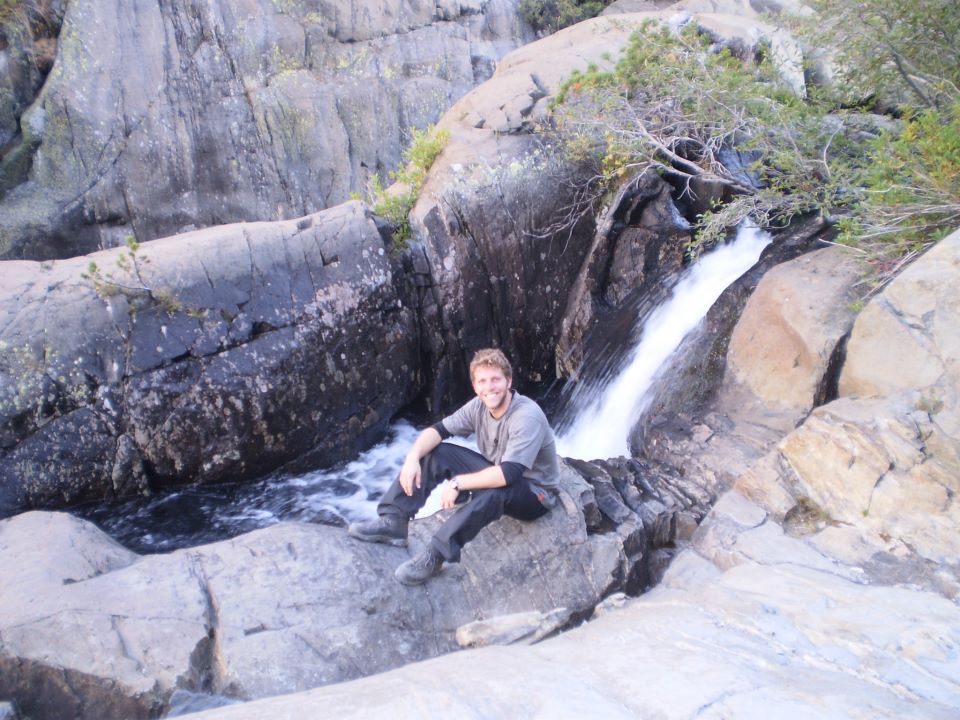 man with arms crossed sitting on large rocks by waterfall