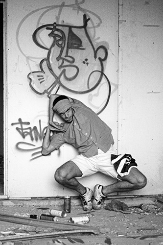 the skateboarder in shorts sits against a wall with graffiti