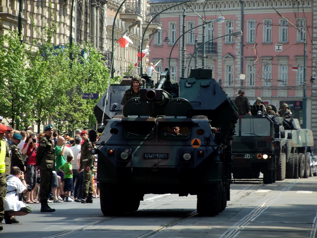 two military trucks drive past a crowd of people