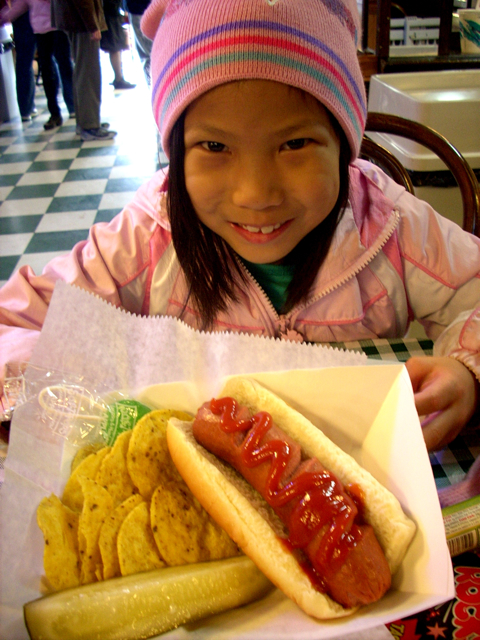 a girl in pink jacket and pink hat sitting at table with dog and some chips