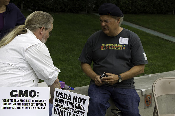 three people sitting at a table talking and one has a sign that reads gmo