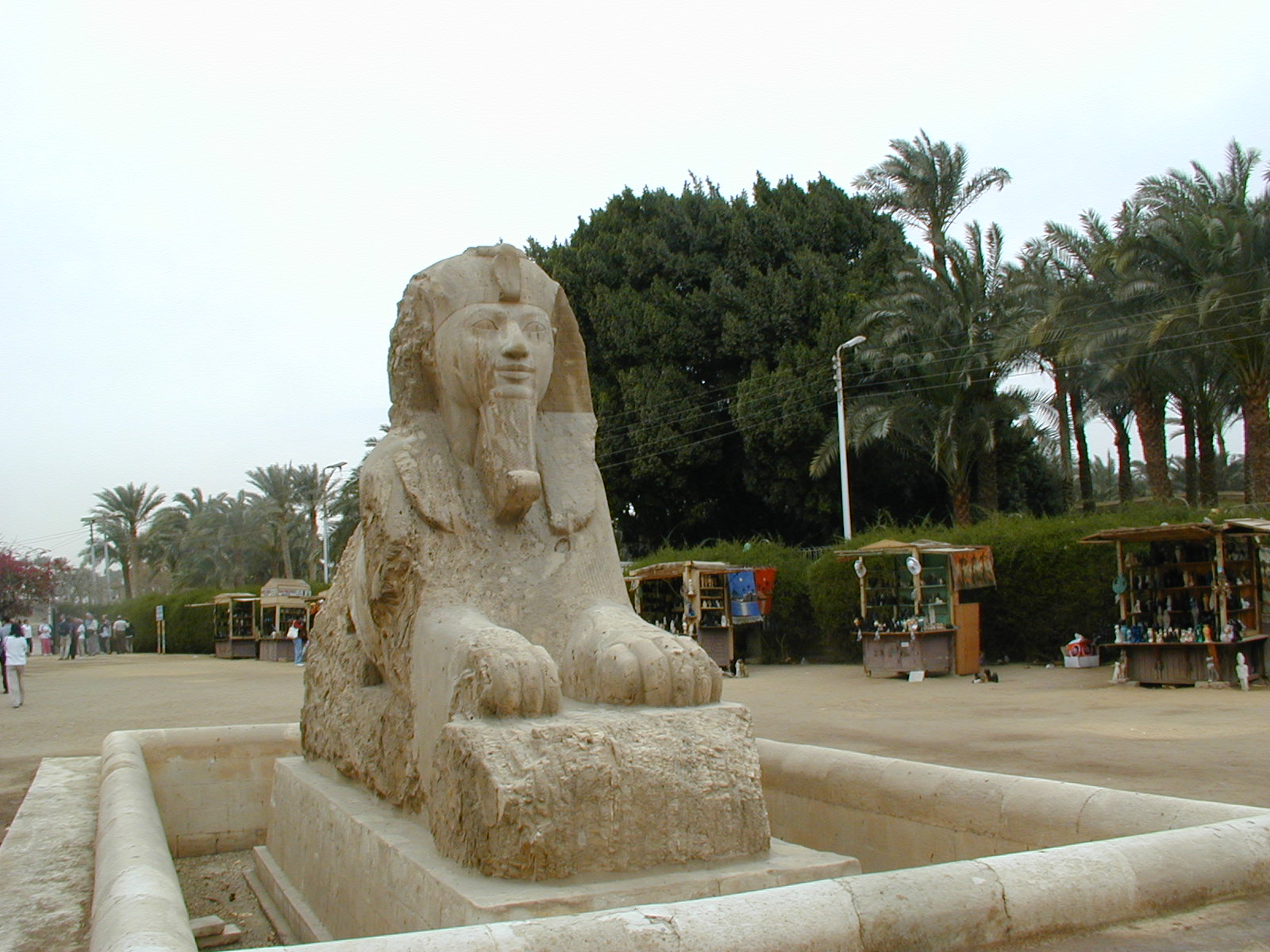 a stone statue of an egyptian king and seated sphinx with palm trees in the background